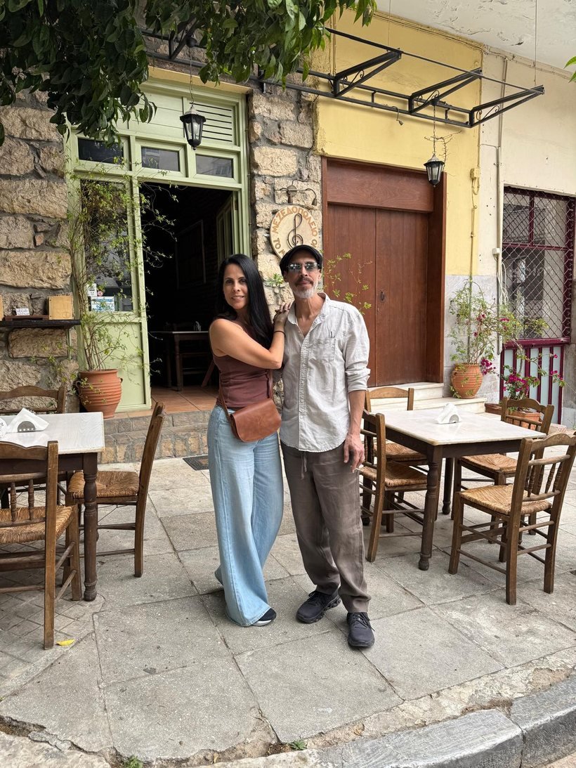 Pnina Havardi with her arm around her brother Roy Havardi, standing outside a stone taverna with a treble-clef sign